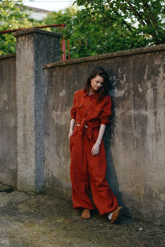 Cheerful woman outdoors, wearing a bold orange jumpsuit, leaning against a weathered wall, relaxed pose, natural light, lush greenery behind, casual lifestyle moment