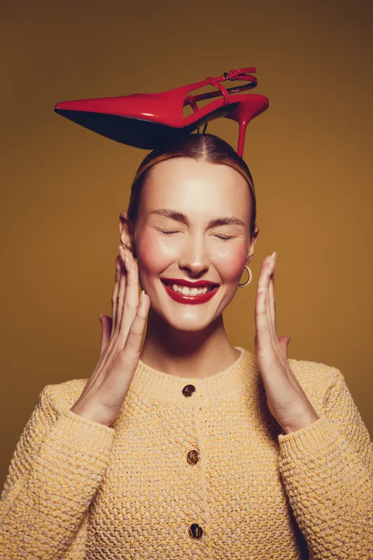 Smiling woman with closed eyes balances red high heel shoe on her head. She wears a yellow textured sweater and gold hoop earrings against a warm brown background. Concept of fun and fashion.