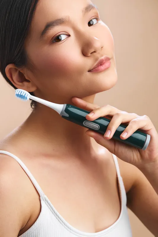 Asian woman holds an electric toothbrush near her face, smiling softly in a bright bathroom. The scene conveys personal care, health, and a calm daily routine in a clean, comfortable setting.