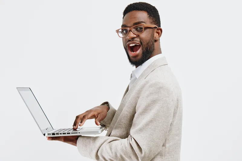 Smiling happy young man with glasses in beige suit holding open laptop looking at camera isolated on plain background. Male facial expression gesture concept