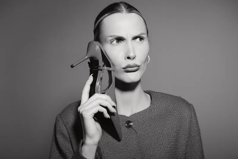 A white woman with light skin holds a high heel shoe to her ear like a phone, showing a confused expression in monochrome.