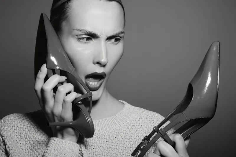Young white woman with light skin looks confused holding two high heel shoes in hands. Studio portrait with fashion concept.