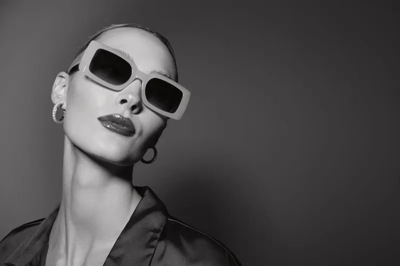 Portrait of a pale white woman wearing large sunglasses and hoop earrings looking confident in studio lighting.