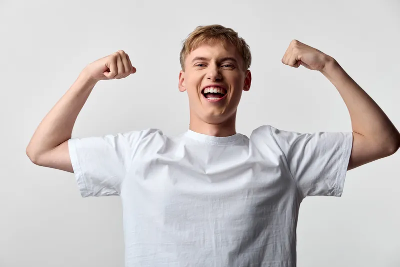 Happy man flexes arms in a victory pose wearing a white t shirt against a plain studio backdrop, exuding confidence, energy and bold motivation for fitness, wellness and celebration themes