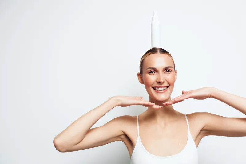 Playful woman balances a bottle on her head, smiling with arms framing her face, wearing a white tank top, in a clean studio, light mood, humorous portrait.