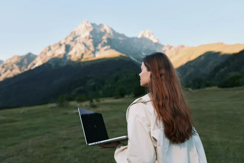 A woman stands in a meadow with a laptop, gazing toward distant mountains under a clear blue sky. She wears a light jacket, focused and calm, embodying remote work in nature.
