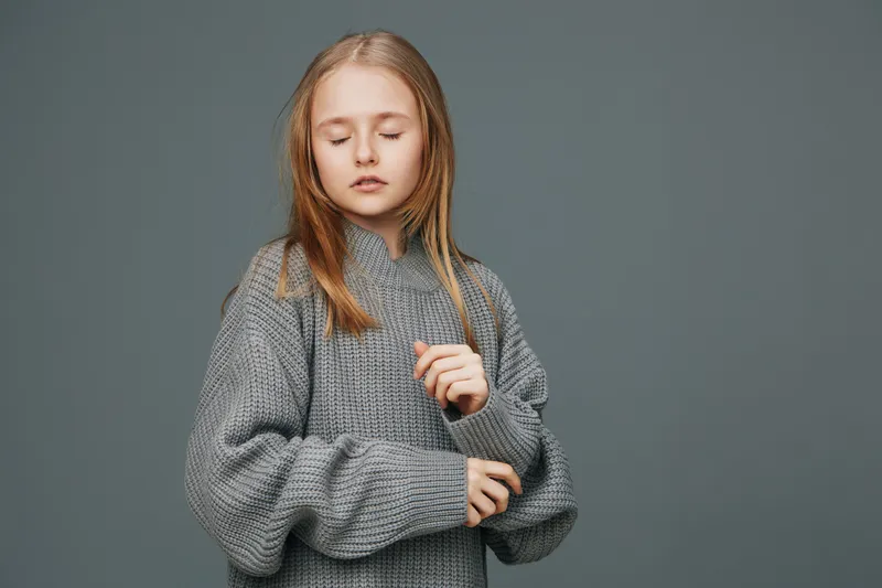 Young girl in a cozy gray sweater, with closed eyes and a serene expression, against a soft gray background, evoking feelings of comfort and calmness