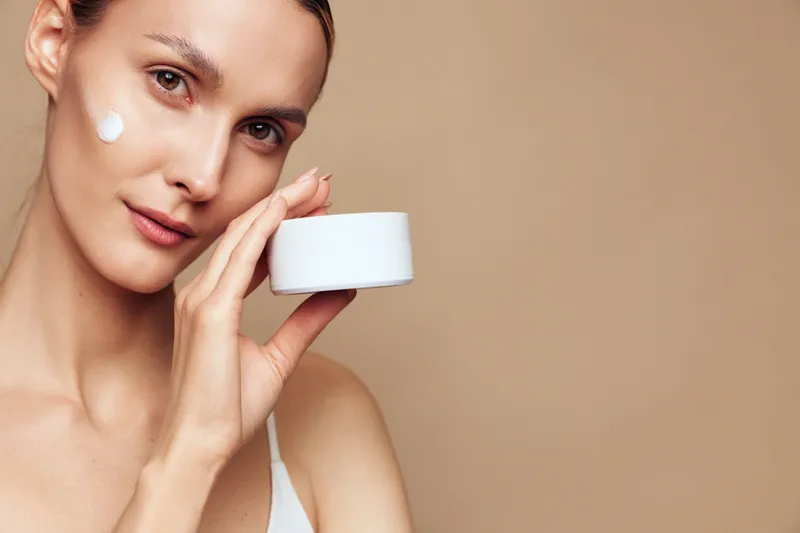 A young adult female with brunette hair applies cream to her cheek in a close up, beige background, capturing a skincare moment and the calm routine, for a fresh, healthy look.