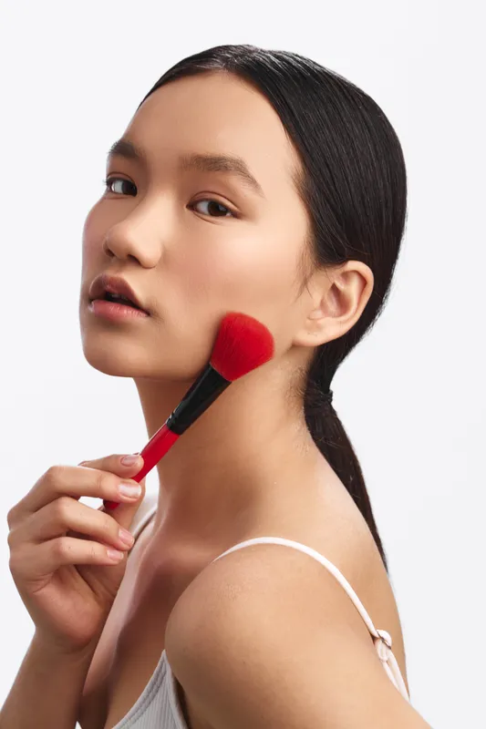 Asian woman applying makeup with a brush in a clean studio setting, showcasing focus and beauty. A soft portrait with gentle lighting highlights skin tone and quiet confidence.