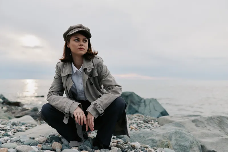 Young woman in casual outfit crouches on rocky beach near calm sea under cloudy sky. Concept of fashion, nature, and thoughtful mood outdoors by the water.