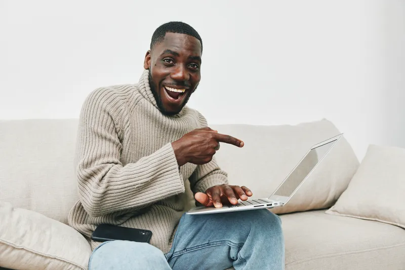 Cheerful young man in a knitted sweater joyfully pointing at his laptop on a cozy sofa, showcasing a welcoming home environment with a light color palette