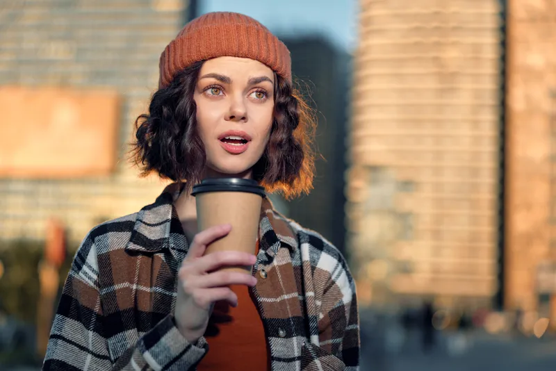 Woman holding coffee in urban street, beanie and plaid jacket at golden hour glow, candid lifestyle portrait with authenticity, mindful living and emotional storytelling in modern city scene.