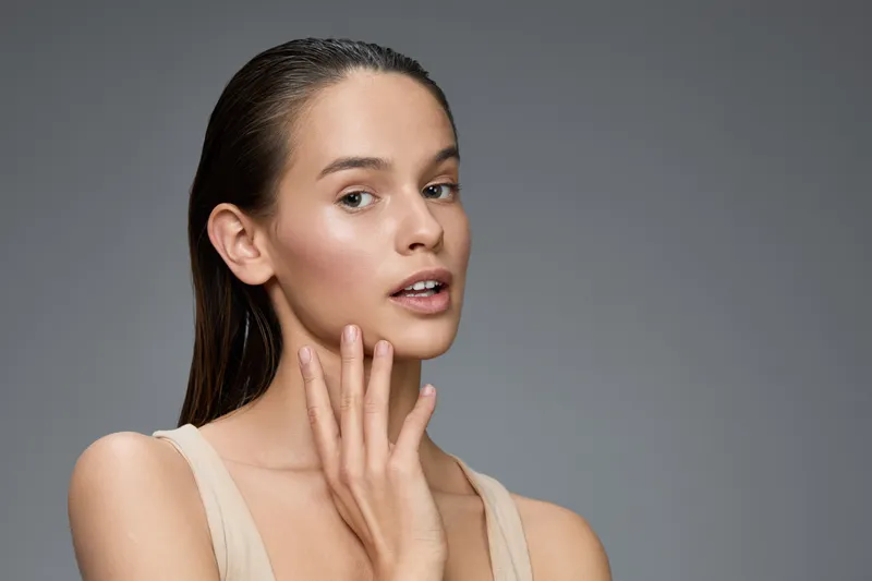 Young woman with glowing skin and natural makeup, expressing confidence against a gray backdrop, ideal for beauty and skincare concepts