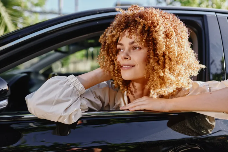 Smiling woman with curly hair leaning out of black car window enjoying sunny day, casual clothing, relaxed mood, natural light, outdoors setting.