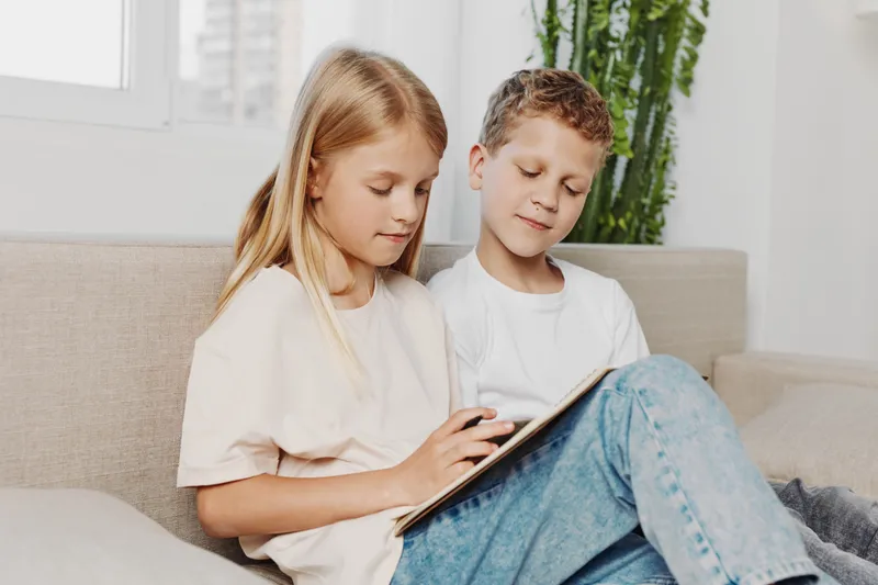 Two children sitting on couch use digital tablet together in bright living room. Casual clothing and relaxed mood show modern family lifestyle and technology use.