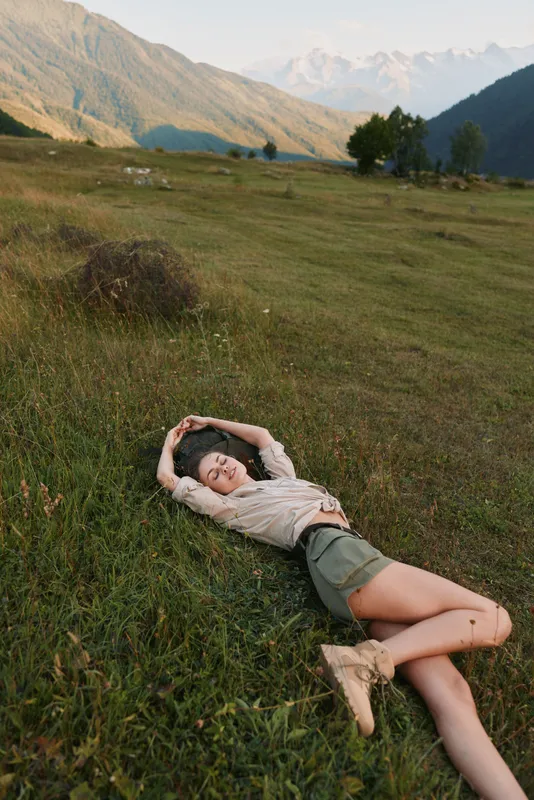Young woman lies on grassy meadow in a mountain valley, hands behind her head, smiling softly as bright daylight and distant peaks frame a serene outdoor portrait.