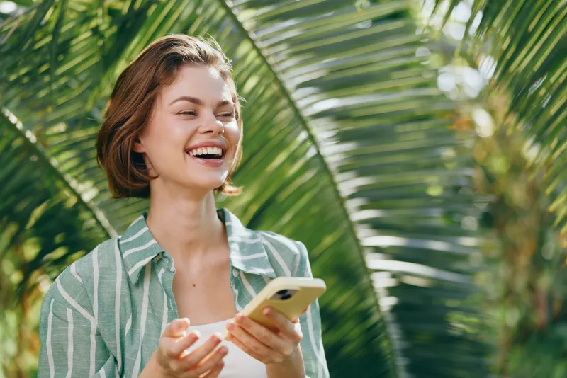 A smiling woman outdoors using a smartphone, surrounded by lush palm leaves and bright natural light.