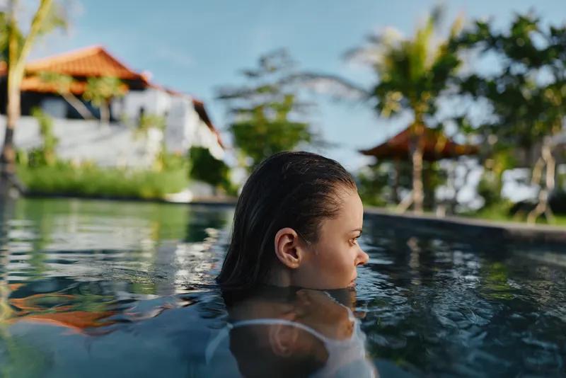 A person relaxes in a pool at a tropical resort, profile view above the water, palm trees in the background. Lifestyle moment emphasizing leisure, vacation, and wellness in a sunny outdoor setting.