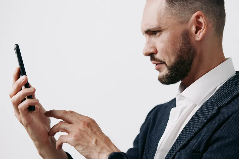 A professional man analyzes data on a smartphone against a solid colored background, conveying focused emotion, confidence, and thoughtful business concentration.