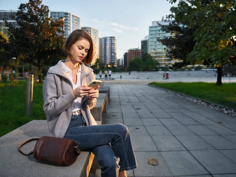 A cheerful woman sits on a bench in a modern city park and texts on her phone, smiling softly as the urban environment surrounds her with calm, friendly energy.