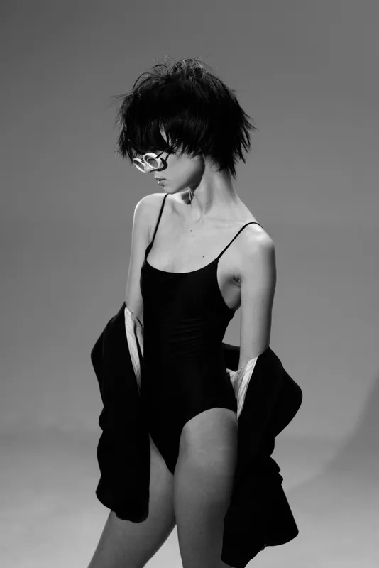 Black and white slender woman in bodysuit with curly hair and glasses posing against gray background