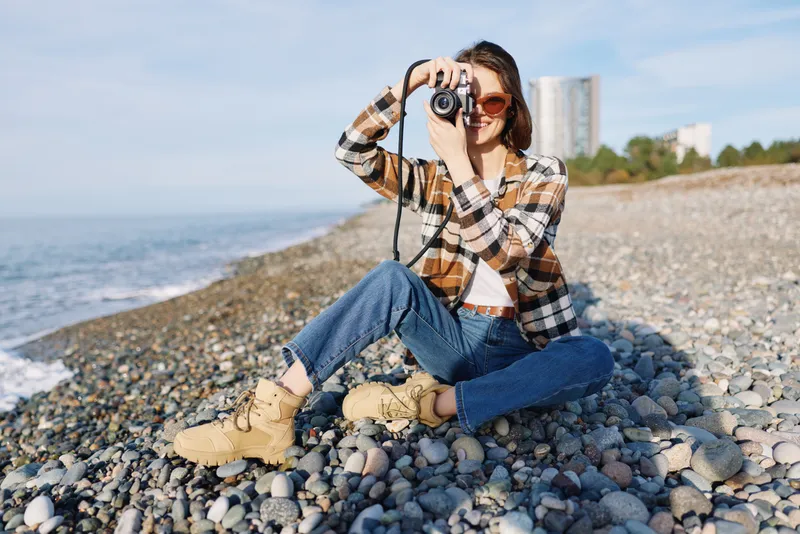 Photographer, woman, beach, camera, outdoors, casual, pebble, coastal, scenery, photography, a woman on a pebble beach crouches with camera to capture coastal scenery on a sunny day, wearing plaid