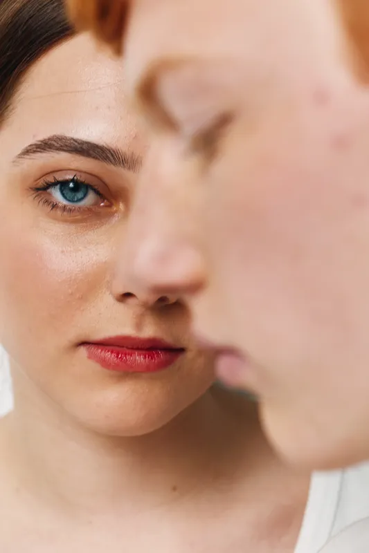 Portrait of woman with blue eyes and red lips close to man face, showing skin texture and natural beauty in soft light on white background.