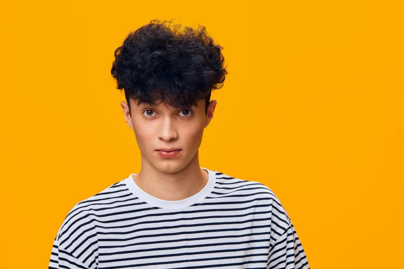 young man with curly hair wearing a striped shirt stands against vibrant orange background with serious expression, studio portrait capturing natural look and casual style