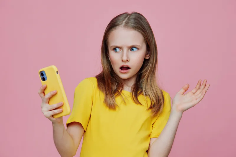 Surprised girl in yellow shirt looking at phone on pink background, expressing shock or disbelief, perfect for youth related concepts and social media themes