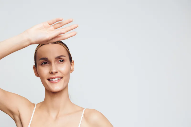Young woman with clear skin smiling and shielding her eyes with her hand. Portrait on a light background. Concept of skincare, beauty, health, and natural wellness.
