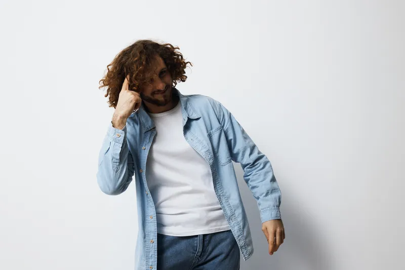 Young man with curly hair in casual blue shirt poses thoughtfully against a plain background, capturing emotions of contemplation and imagination
