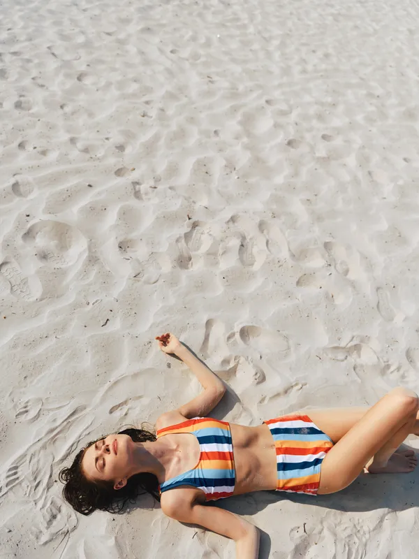 A young woman lies on sunlit beach sand in a colorful striped bikini, relaxed pose and sunny glow, embodying carefree summer leisure and playful vacation vibes.