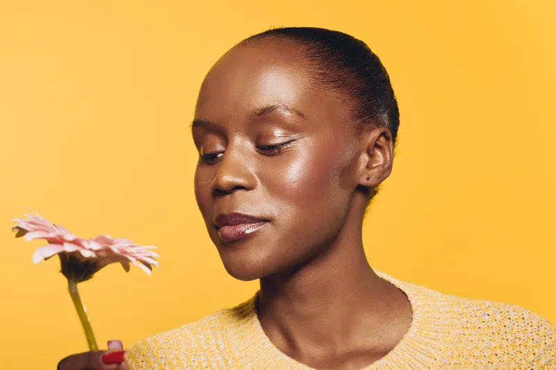 Close-up of a dark skinned African woman with smooth skin holding a pink flower near her face against a yellow background. Concept of natural beauty, skincare, and self-care.