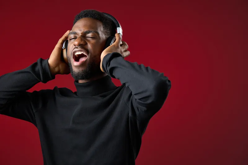 A young African man joyfully singing while wearing headphones against a vibrant red background, showcasing emotions of happiness and enthusiasm