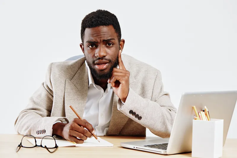 Thoughtful young african american man in formal beige blazer and white shirt sitting at office desk with laptop and glasses, appearing to be in deep concentration or reflection, isolated on plain whit