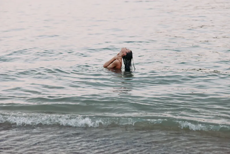 A solitary swimmer stands waist deep in a peaceful sea, fingers brushing the surface while soft waves roll toward a quiet, sandy shore, capturing reflective morning light.
