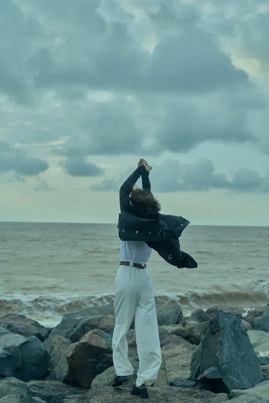 Man raises a jacket in the windy seashore, clothes billowing, capturing motion and freedom against the ocean and cloudy sky. Wide horizon. Dramatic mood.