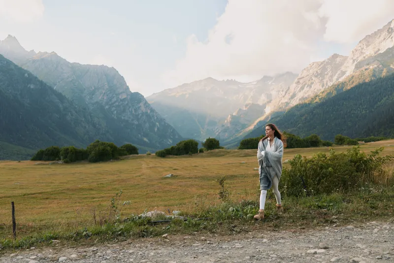 Young woman walks along a gravel road through a mountain valley, wearing a denim jacket and scarf, enjoying the wide landscape and fresh air as distant peaks loom