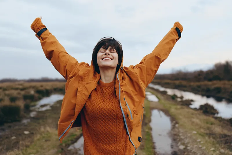 Woman lifestyle outdoors on a hiking trail, smiling with arms raised in an orange jacket, enjoying nature beside a river, scenic sky and countryside background