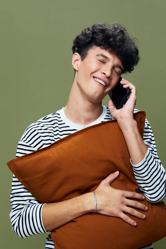 Smiling man holding brown pillow and talking on phone, wearing striped shirt with curly hair on green background, expressing joy and relaxation in casual style.