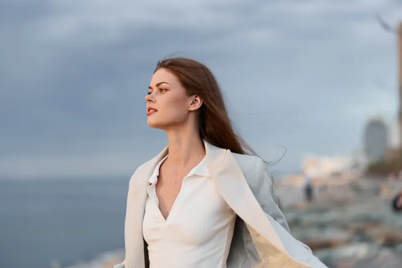 Confident woman in white sweater and beige coat standing by the sea with serene expression during cloudy weather blue sky background autumn fashion style