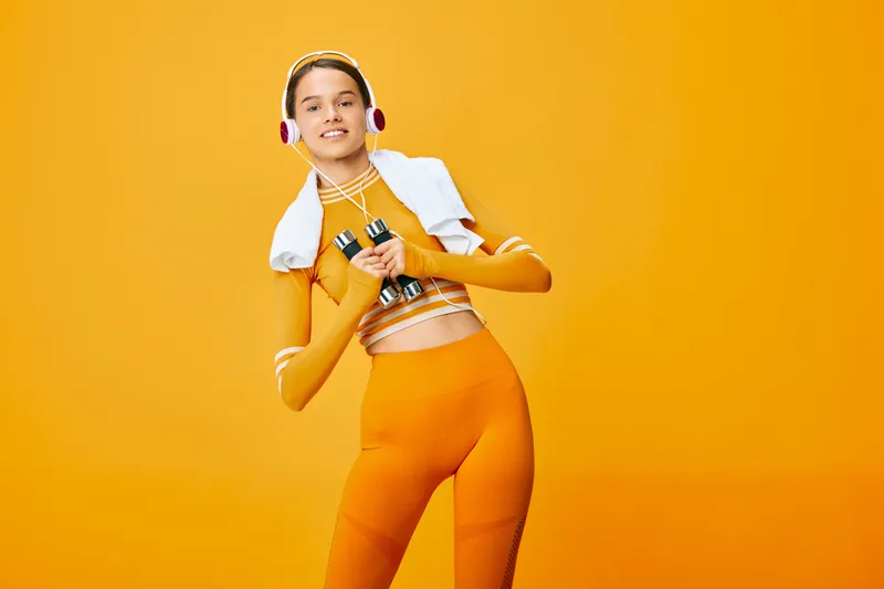 Fit woman in orange activewear holds dumbbells and wears headphones with a white towel draped over her shoulders against a vivid orange studio background, smiling confidently.