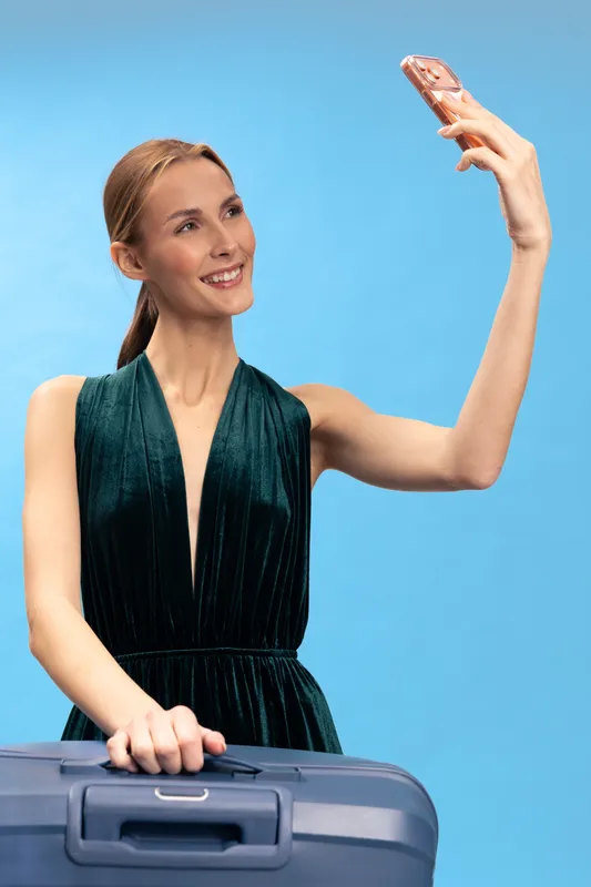 Young smiling woman in dark green dress holds suitcase and takes selfie with smartphone on blue background. Concept of travel, vacation, happiness, and modern communication.