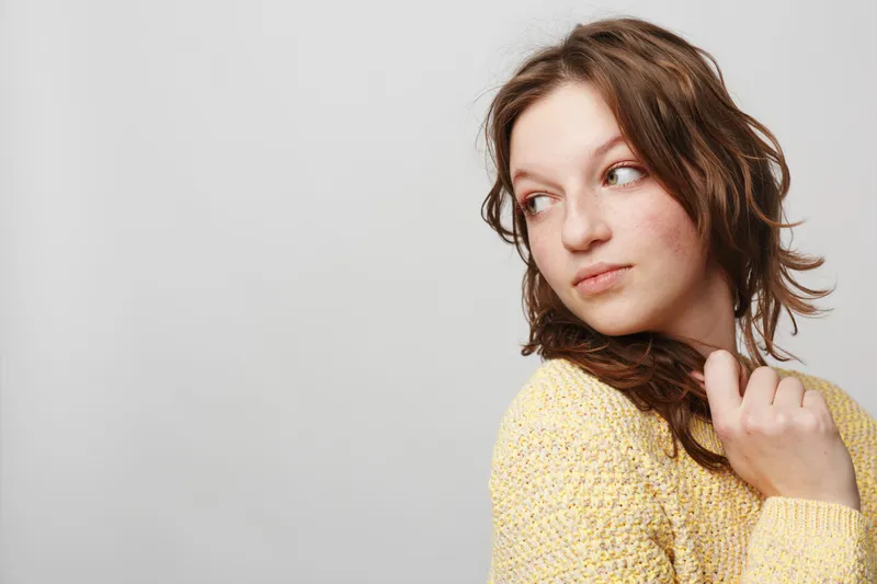 Young woman with wet hair wearing yellow knitted sweater looks sideways with thoughtful expression on light grey background. Concept of contemplation, casual style, and natural beauty.