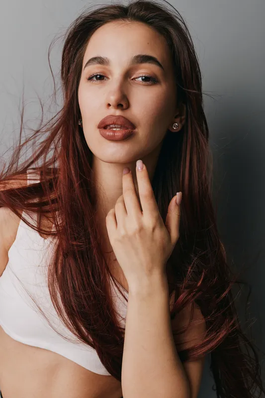 Young woman with long brown hair posing with hand near face, wearing white top, natural makeup, and earrings in studio with neutral background and soft lighting.