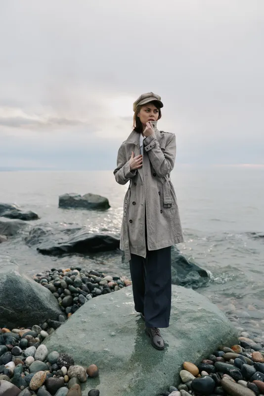 A woman in a patterned coat and cap stands on a rocky shore near calm water under a cloudy sky. The image conveys fashion, solitude, and coastal nature elements.