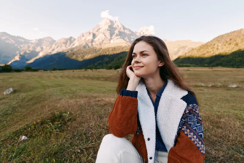 Woman smile mountains meadow portrait of a young outdoor scene with cozy jacket and sunlight, serene nature landscape capturing thoughtful relaxation, travel mood and peaceful beauty.