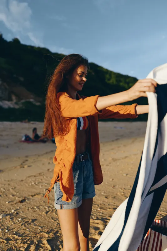 A young woman with long curly hair wearing an orange shirt and denim shorts spreads a towel on a sandy beach during a sunny day with hills in the background.