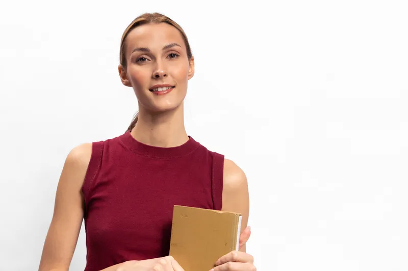 Young woman in sleeveless burgundy top holds a book and smiles at camera. Studio portrait on white background with natural lighting. Concept for education, reading, and knowledge.