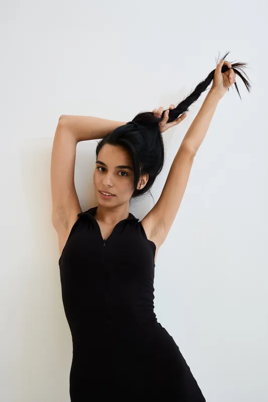 Young woman in black sleeveless dress holding her long braided hair up against white background, showcasing natural beauty and confident pose.